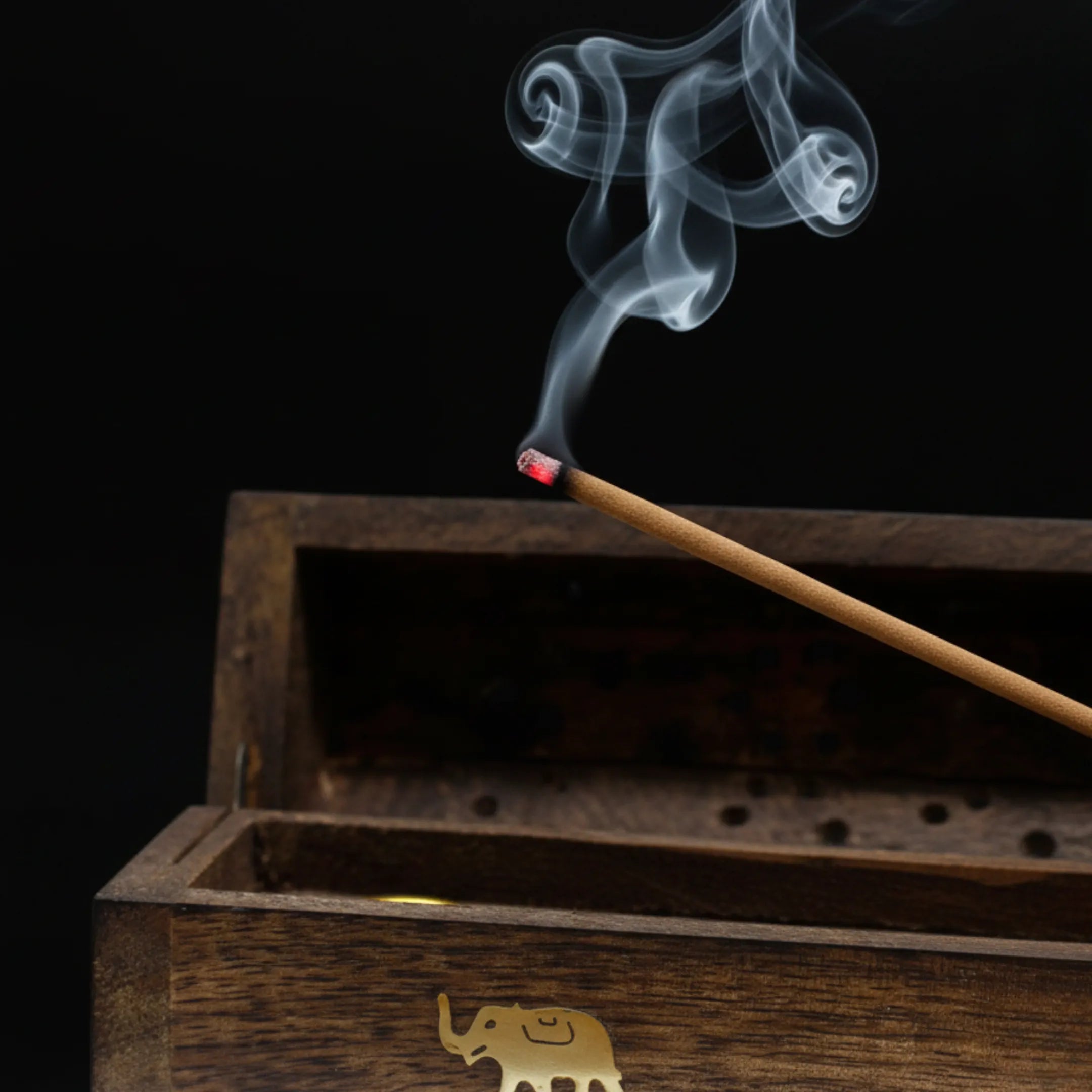 Zoom macro sur un bâtonnet d'encens qui est en train de se consumer, posé dans une boîte support d'encens en bois le tout sur fond noir.