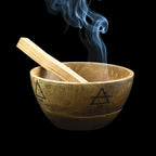 Bol en bois de manguier, symboles des 4 éléments gravés, avec bâton de Palo Santo allumé à l'intérieur, sur fond noir.
