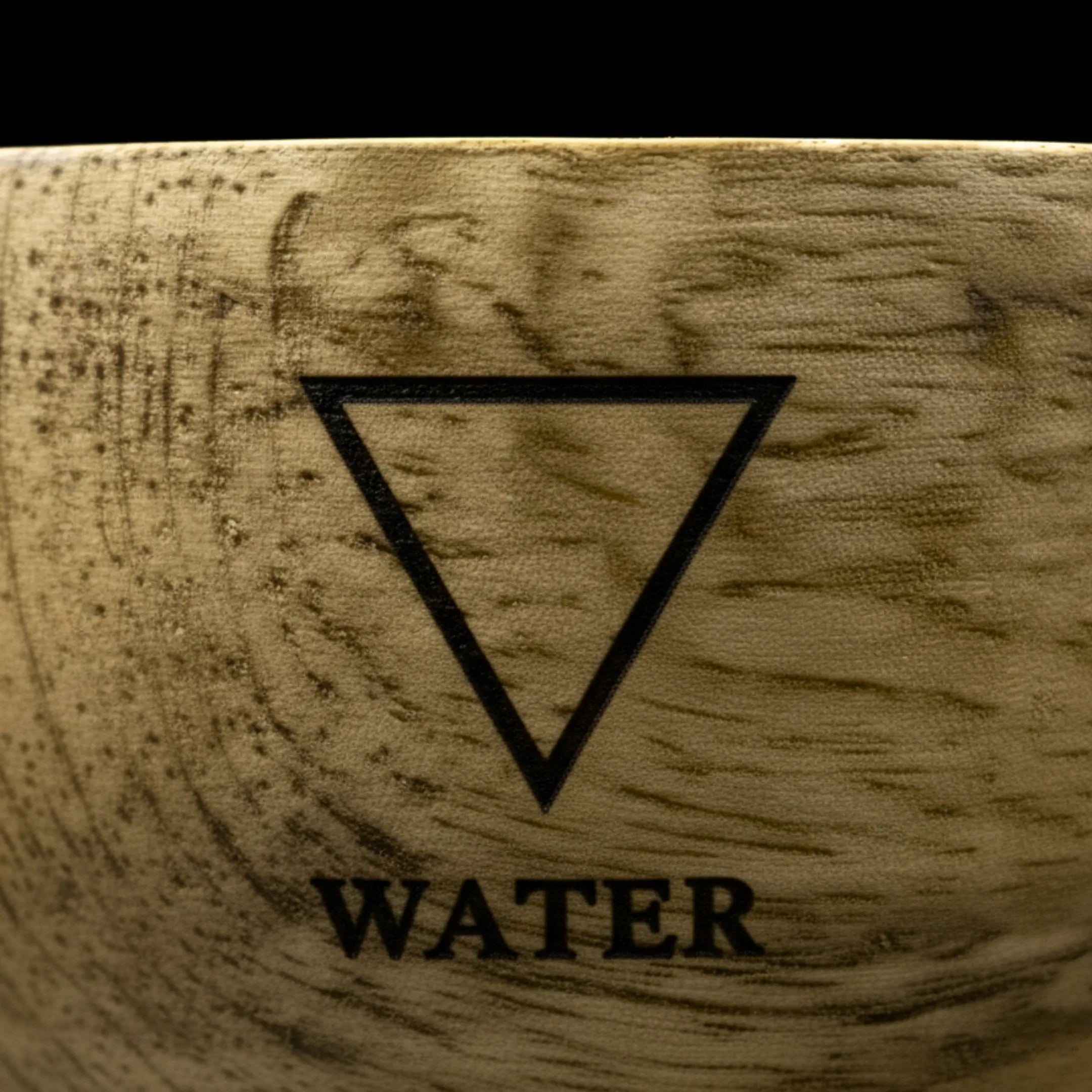 Bol en bois de manguier avec un zoom macro sur le symbole de l'élément eau, gravé sur le haut du bol, sur fond noir.
