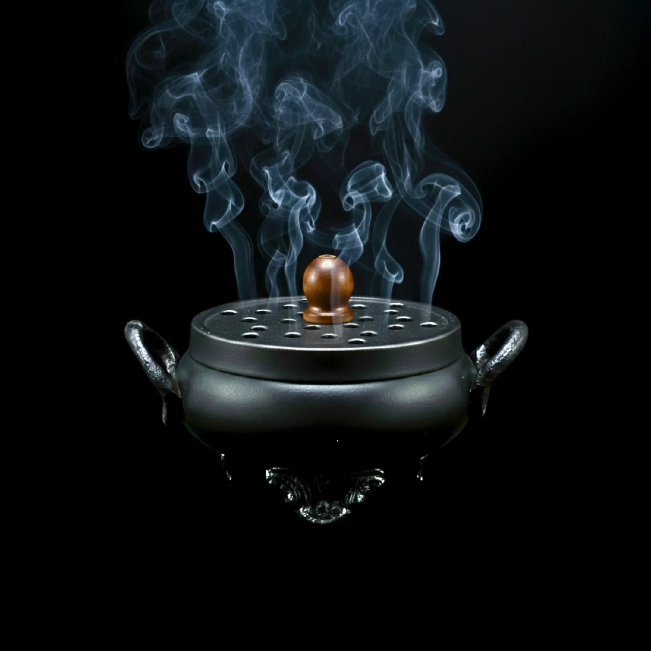 Brûleur à encens chaudron en fonte avec couvercle et poignée en bois, la fumée sortant des trous, sur fond noir.