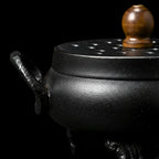Brûleur d'encens en fonte, en vue macro, en forme de chaudron, avec couvercle et poignée en bois, sur fond noir.
